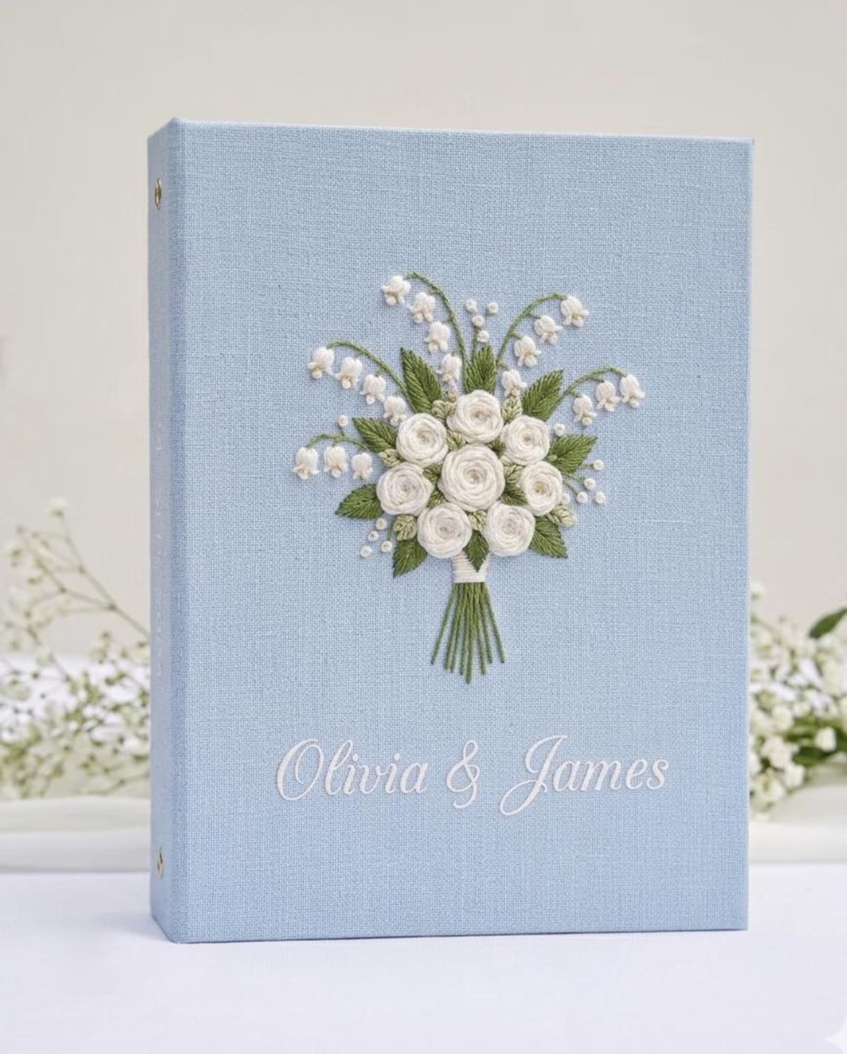 A light blue book with embroidered white roses and lily of the valley — hand-embroidered by The Thread Nest