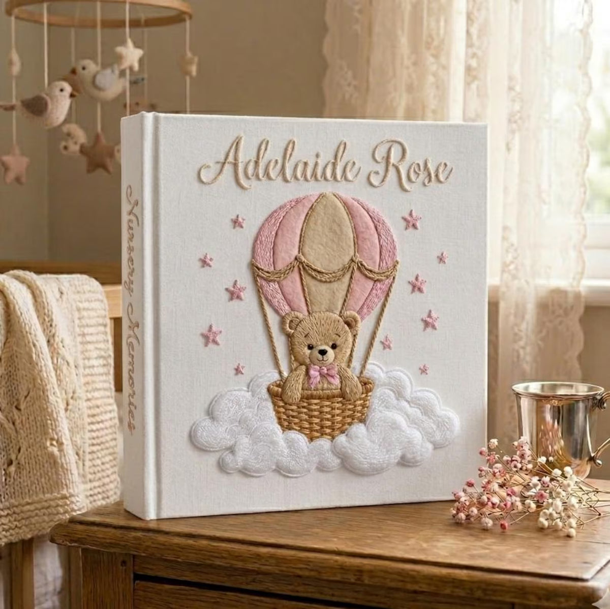 A white memory book with a teddy bear in a tan and pink hot air balloon — hand-embroidered by The Thread Nest