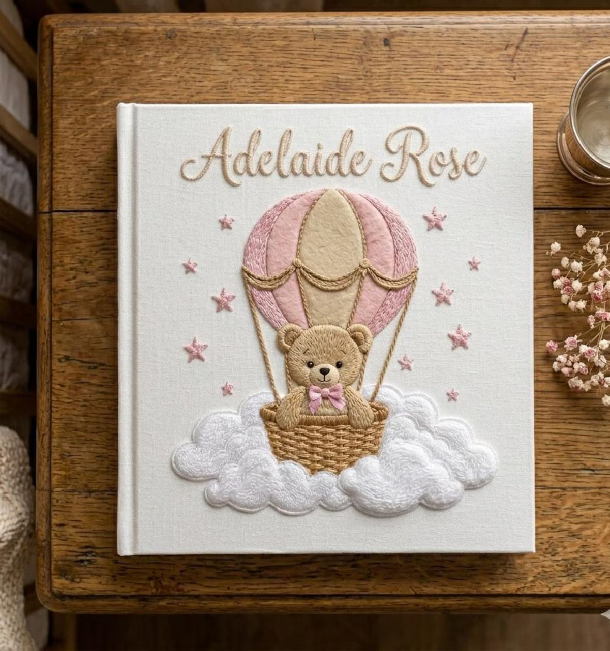 A square white book with a tan teddy bear in a bow and a stitched name — hand-embroidered by The Thread Nest