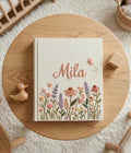 A cream-colored book with a name in pink script, flowers and butterflies — hand-embroidered by The Thread Nest