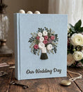 A light blue wedding guest book with an embroidered floral bouquet design in white, pink, and purple — hand-embroidered by The Thread Nest