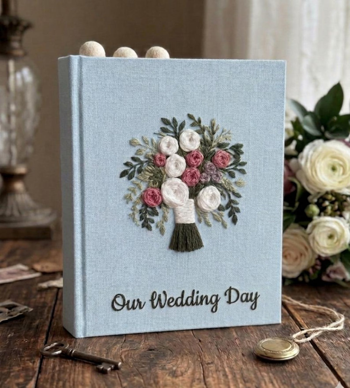 A light blue wedding guest book with an embroidered floral bouquet design in white, pink, and purple — hand-embroidered by The Thread Nest