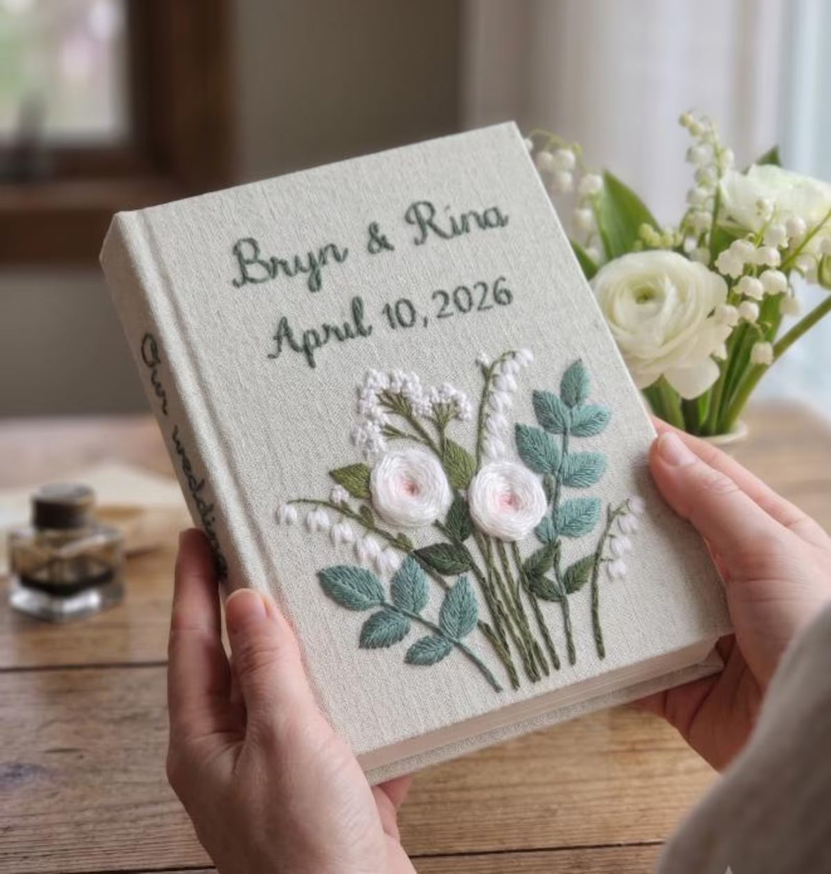 A light beige book with embroidered names, a date, and Our wedding on the spine — hand-embroidered by The Thread Nest