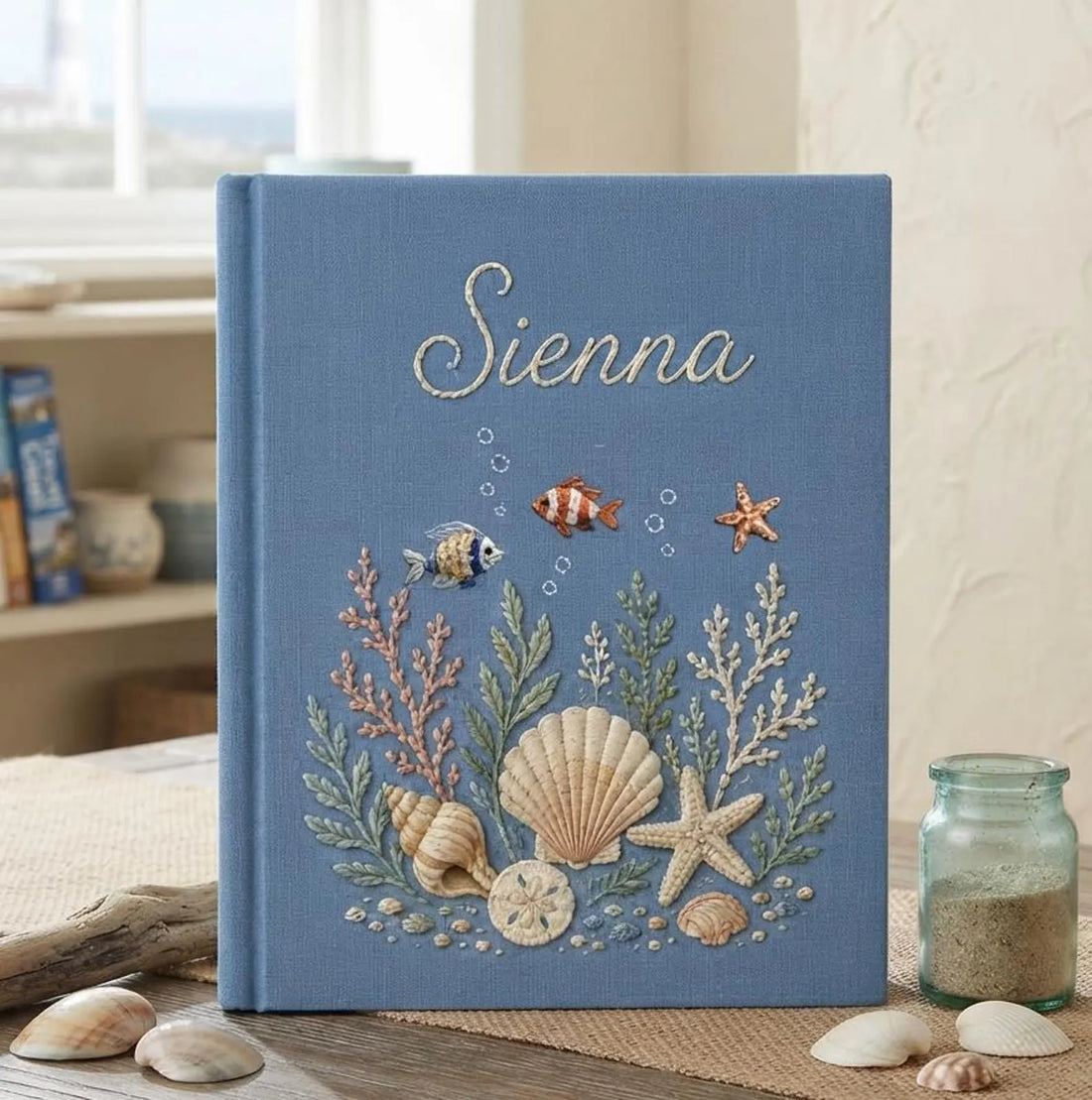 A blue book with an embroidered underwater scene of seashells and a stitched name — hand-embroidered by The Thread Nest