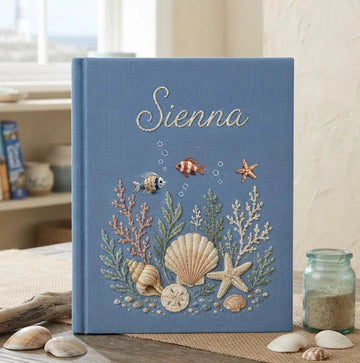 A blue book with an embroidered underwater scene of seashells and a stitched name — hand-embroidered by The Thread Nest