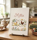 A cream-colored book with embroidered chickens and a name in cursive script — hand-embroidered by The Thread Nest
