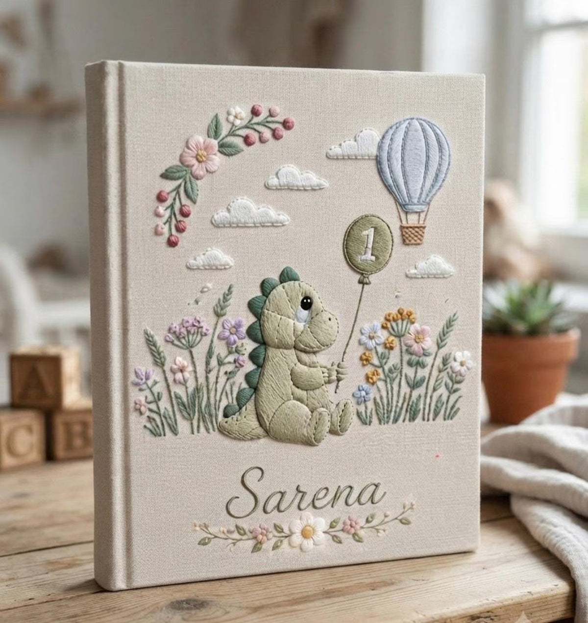A beige book with a green dinosaur holding a number one balloon — hand-embroidered by The Thread Nest
