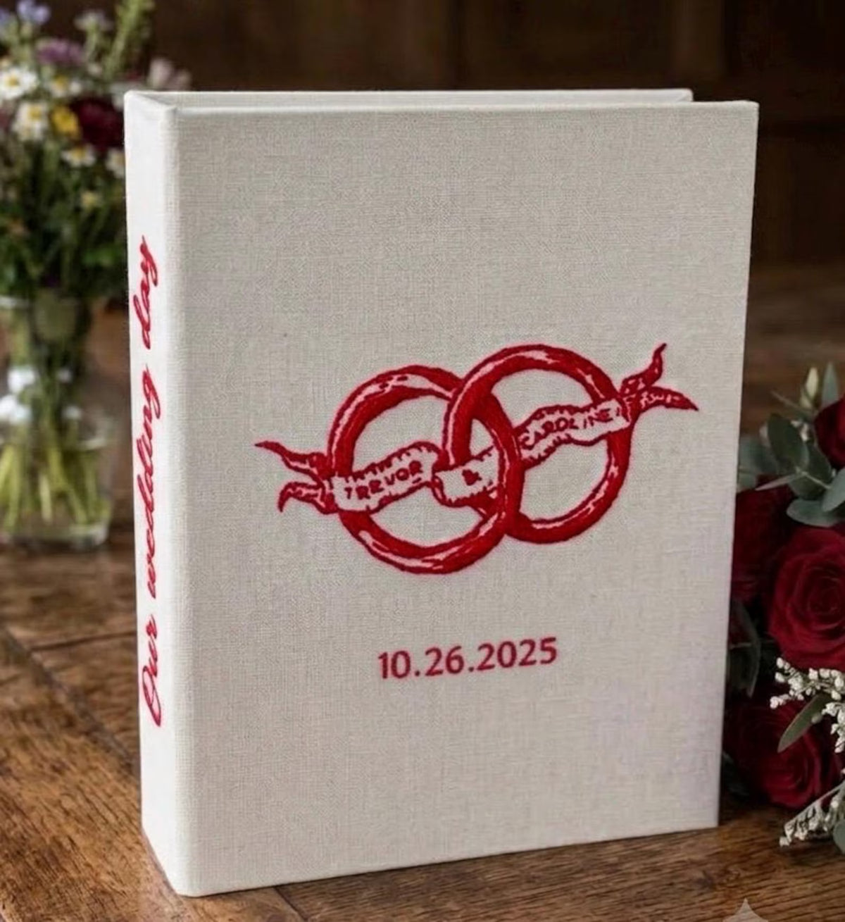 A cream-colored book with red embroidered rings and Our wedding day on the spine — hand-embroidered by The Thread Nest
